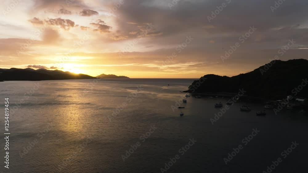 Sunset over Junquillal Bay in Costa Rica. Silhouetted mountains, golden sunbeams piercing through clouds. Small boats dot the calm waters, shimmering reflections across ocean surface.