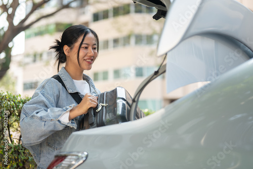 A smiling Asian woman places her luggage in the trunk of a car, getting ready for her trip. photo