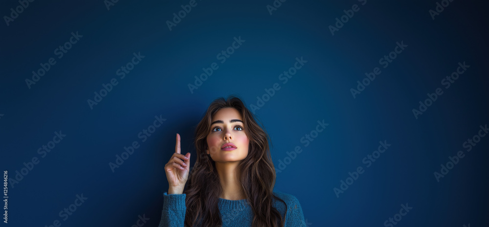 A young woman looks upward and points an index finger set against a dark blue background. for words and banner A young woman looks upward and points an index finger set against a dark blue background. for words and banner