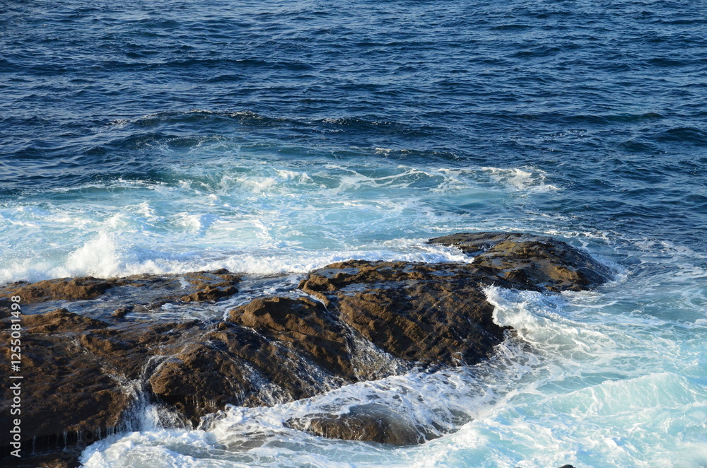 Fototapeta premium Waves crashing on dark rocks along the Baiona coastline in Galicia, blending blue ocean hues with white foam, capturing the raw energy of the Atlantic 