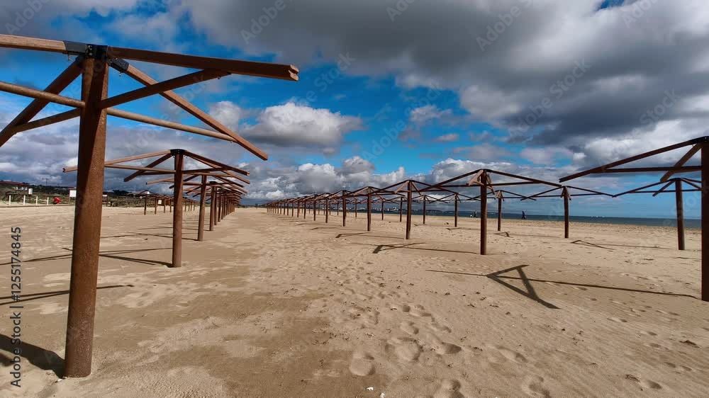 A long row of empty beach umbrellas stretches along a sandy beach. The sky is a bright blue with fluffy white clouds. In Lagos, meia praia, Portugal. A long row of empty beach umbrellas stretches along a sandy beach. The sky is a bright blue with fluffy white clouds. In Lagos, meia praia, Portugal.
