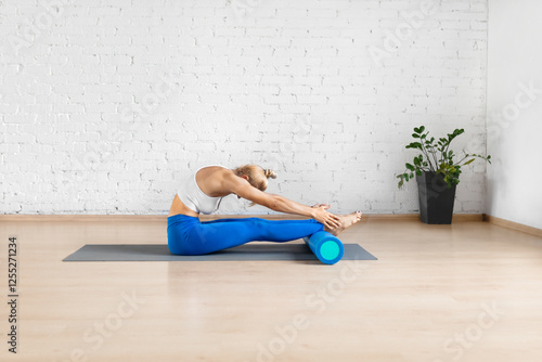 Pilates workout with props. Fit woman does seated forward bend exercise using a foam roller under her feet in fitness studio indoors. photo