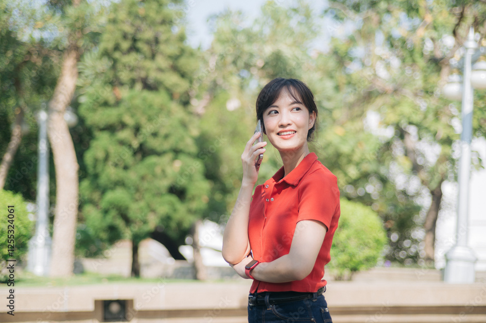 Asian cheerful young woman in red shirt talking on her phone while standing outdoors in sunny park Asian cheerful young woman in red shirt talking on her phone while standing outdoors in sunny park