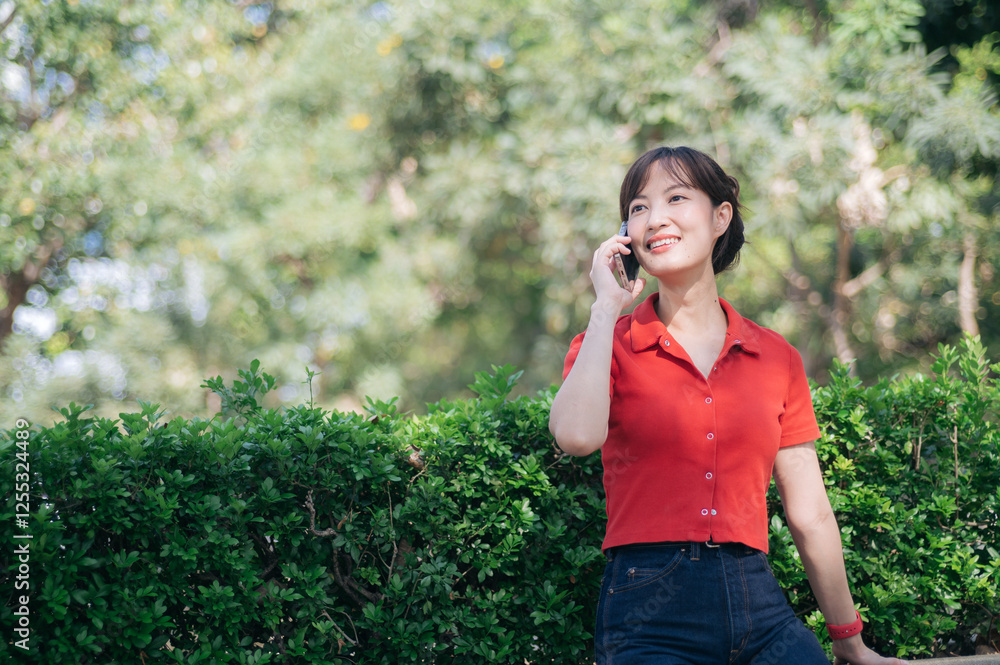 Asian cheerful woman in red shirt talks on her phone while standing near green hedge in sunny outdoor park Asian cheerful woman in red shirt talks on her phone while standing near green hedge in sunny outdoor park