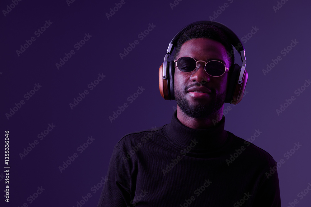 Obraz premium Confident young man wearing sunglasses and headphones, posing against a purple background, showcasing modern style and urban fashion, perfect for music and lifestyle themes