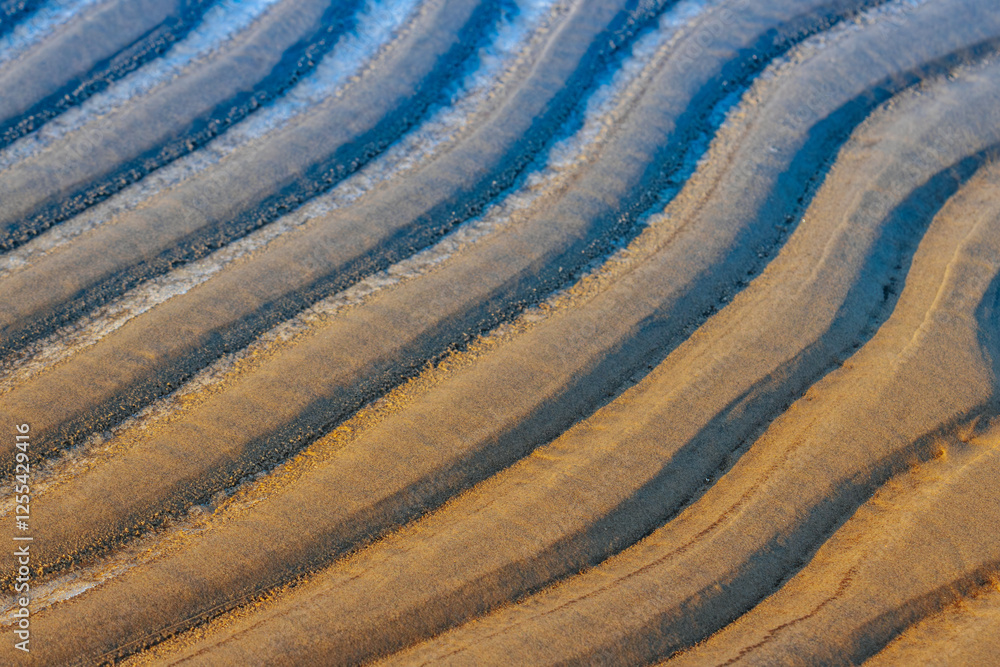 Close-up of wet sand ripples with dark and light patterns formed by receding water, creating a textured and natural abstract design. A mix of golden, brown, and blue tones enhances the contrast. Close-up of wet sand ripples with dark and light patterns formed by receding water, creating a textured and natural abstract design. A mix of golden, brown, and blue tones enhances the contrast.