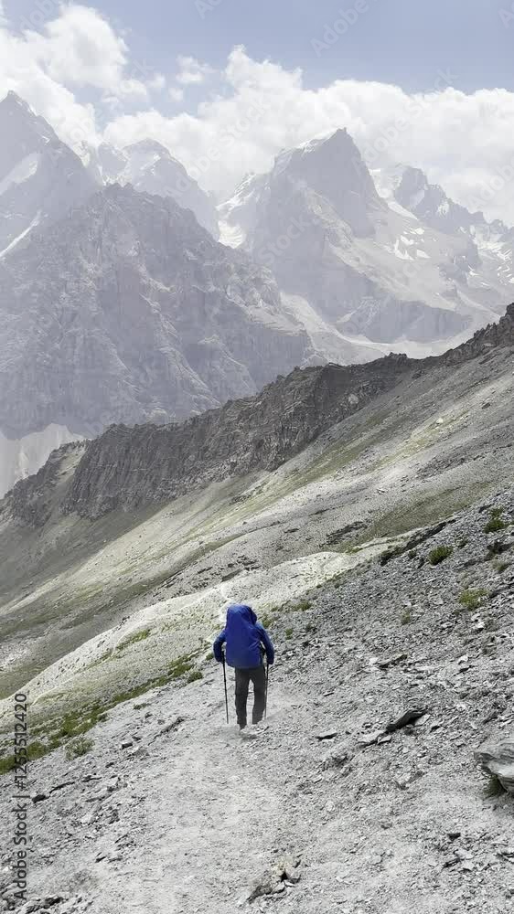 A young man with a hiking backpack walks down a mountain path overlooking snowy mountain peaks. Trekking in the mountains , summer travel and outdoor activities A young man with a hiking backpack walks down a mountain path overlooking snowy mountain peaks. Trekking in the mountains , summer travel and outdoor activities