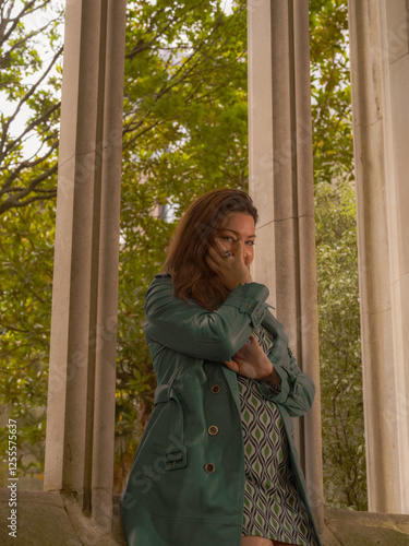 A woman in a blue coat, patterned dress, and matching boots poses playfully on cobblestone ground in front of an old stone window frame with greenery