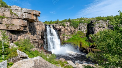Full outdoor shot of a waterfall cascading down rocky cliffs amidst lush greenery under a vibrant blue sky. The water sprays outward from the falls