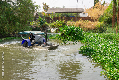 Water hyacinth removal boat operating in a canal in Thailand, using a mechanical arm to extract aquatic plants, helping to prevent water pollution and maintain a clean, navigable waterway in a rural