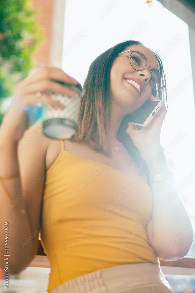 Cheerful hipster girl in classic spectacles enjoying positive cellphone calling during morning coffee time in city, backlit of happy millennial female tourist with takeaway cup laughing while talking Cheerful hipster girl in classic spectacles enjoying positive cellphone calling during morning coffee time in city, backlit of happy millennial female tourist with takeaway cup laughing while talking