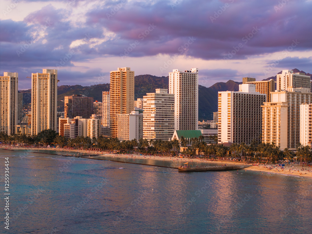 Obraz premium Waikiki Beach in Honolulu, Oahu, Hawaii, at sunset with calm ocean waters, palm lined shore, high rise buildings, and distant mountainous terrain.