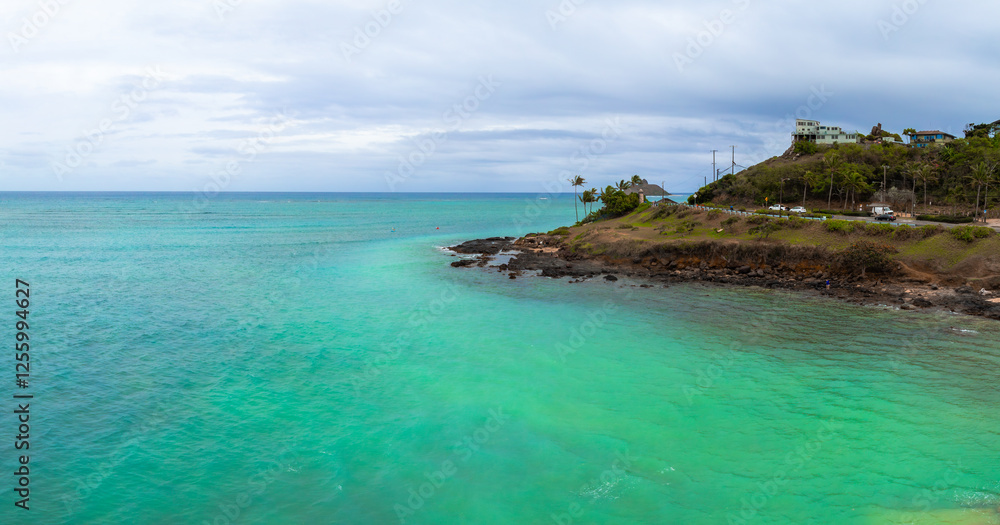 Turquoise waters meet a rocky shoreline on Oahu, Hawaii. A small hill with houses, palm trees, and a road with cars is visible under a cloudy sky. Turquoise waters meet a rocky shoreline on Oahu, Hawaii. A small hill with houses, palm trees, and a road with cars is visible under a cloudy sky.