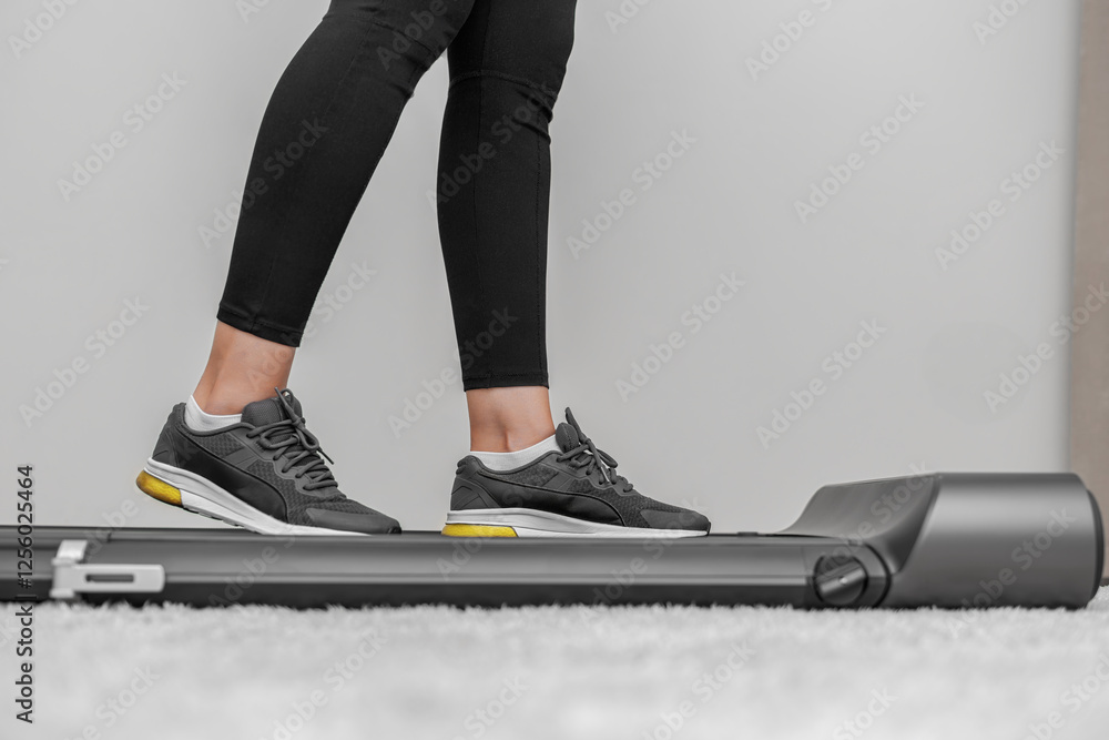 A person wearing black leggings and gray sneakers is walking on a compact treadmill in a modern, minimalistic home environment, focusing on fitness and an active lifestyle.