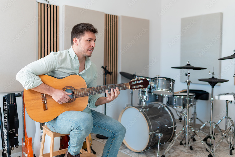 Fototapeta premium A young man playing an acoustic guitar in a professional recording studio with drums and soundproof panels in the background