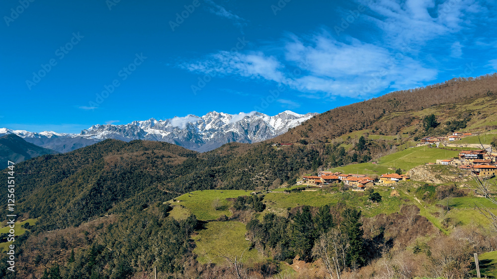 Obraz premium A charming rural village with red-tiled roofs nestled in lush green hills, set against the majestic snow-capped Picos de Europa under a clear blue sky. A peaceful, breathtaking view.