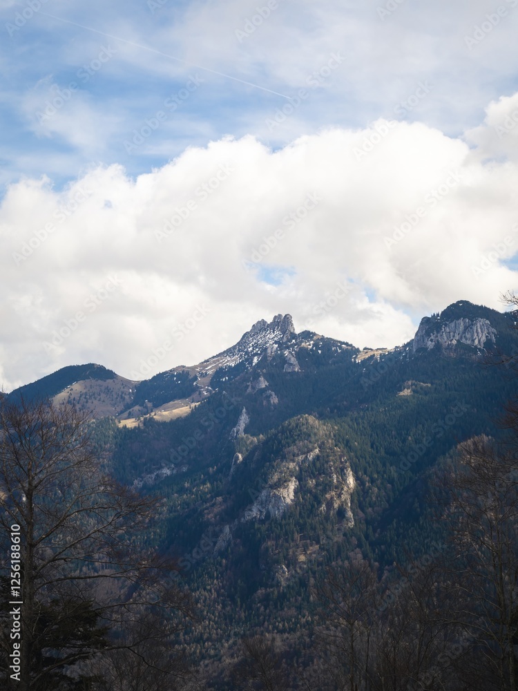Fototapeta premium The Kampenwand mountain range in Bavaria, Germany, under a vibrant sky. Snow-dusted peaks rise above lush forested slopes, creating a breathtaking alpine view.