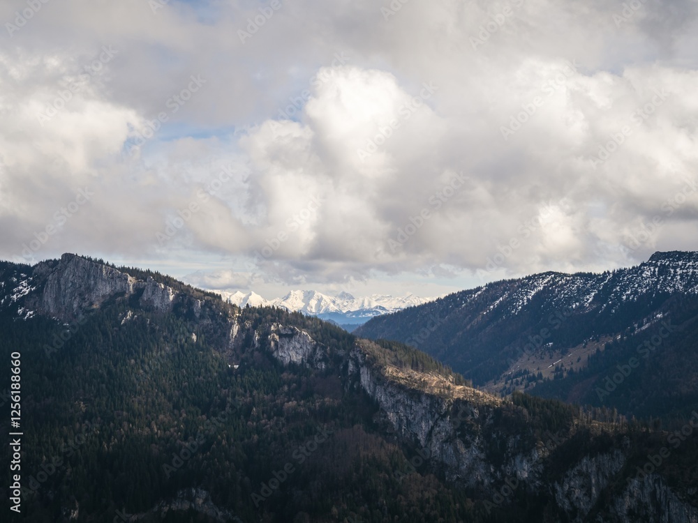 A scenic Bavarian view of a forested mountain ridge with patches of snow, framed by a dramatic cloudy sky and distant snow-capped peaks in the background. A scenic Bavarian view of a forested mountain ridge with patches of snow, framed by a dramatic cloudy sky and distant snow-capped peaks in the background.