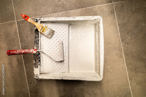 painting tools lying in a tray with white paint. View from above, against a background of gray tiles. A brush and roller, also stained with paint, can be seen in the tray flat lay and copy space. photo