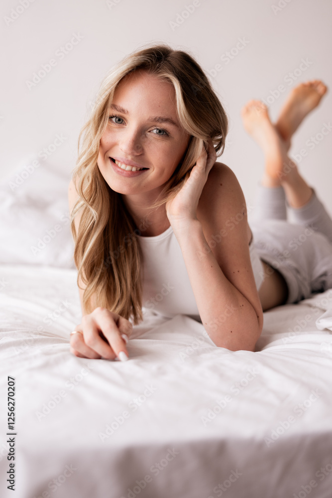 Young happy woman has toothy smile lies on stomach in bed looking at camera spends morning in bedroom Young happy woman has toothy smile lies on stomach in bed looking at camera spends morning in bedroom