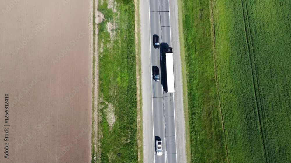 From height of bird flight, picturesque view of two -lane road, which runs through green fields, opens. Sunny rays play on asphalt, creating game of shadows and light. Cargo and passenger cars. From height of bird flight, picturesque view of two -lane road, which runs through green fields, opens. Sunny rays play on asphalt, creating game of shadows and light. Cargo and passenger cars.