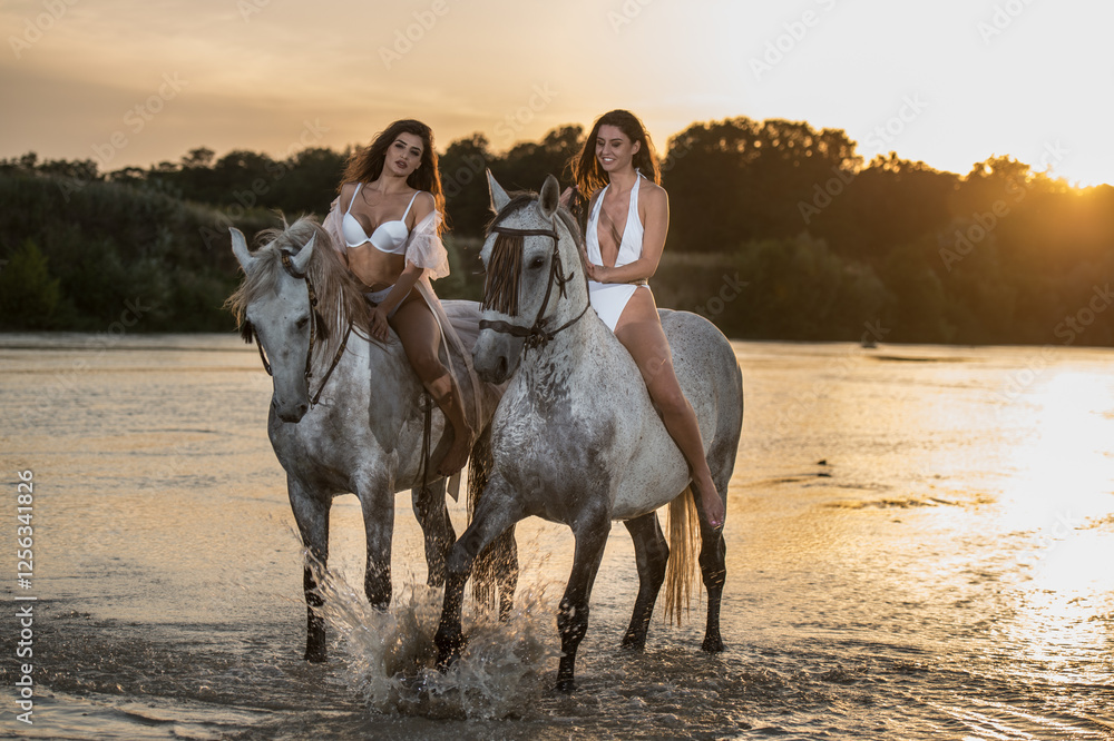 Beautiful brunettes woman with long hair ,white bikini and white shirt rides on a white horse in the river. Sensual and attractive girls interacting and rides a horse in the water at sunset