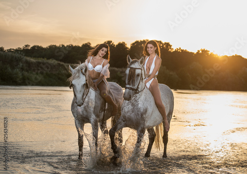 Beautiful brunettes woman with long hair ,white bikini and white shirt rides  on a white horse in the river. Sensual and attractive girls  interacting and rides a horse in the water at sunset