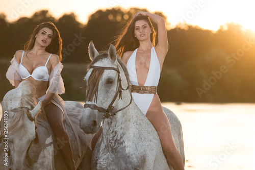 Obraz na płótnie Beautiful brunettes woman with long hair ,white bikini and white shirt rides on a white horse in the river. Sensual and attractive girls interacting and rides a horse in the water at sunset w salonie