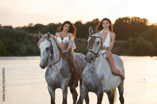 Obraz na płótnie Beautiful brunettes woman with long hair ,white bikini and white shirt rides on a white horse in the river. Sensual and attractive girls interacting and rides a horse in the water at sunset w salonie
