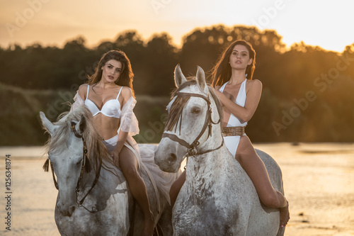 Beautiful brunettes woman with long hair ,white bikini and white shirt rides  on a white horse in the river. Sensual and attractive girls  interacting and rides a horse in the water at sunset
