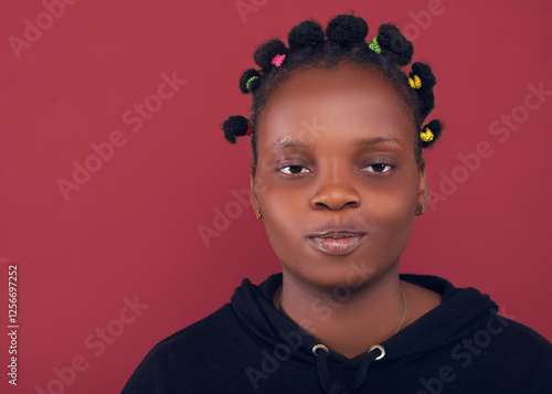 Portrait of a young woman with colorful Bantu knots, wearing a black hoodie, smiling confidently against a burgundy background. Studio lighting highlights her natural beauty and expression. photo