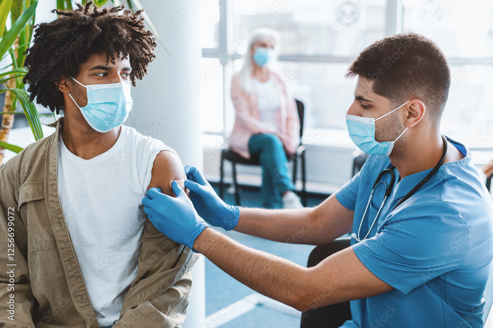 Obraz premium Man doctor putting medical plaster to African American guy patient's arm in clinic office after vaccination injection against covid-19 during pandemic. Vaccinating adults against corona virus concept