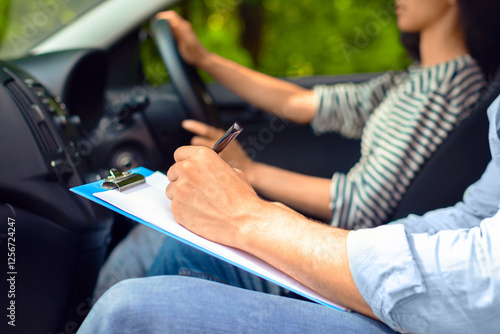 Cropped of male instructor examinating female student, taking notes at test chart while sitting by young woman driving automobile, side view, copy space, closeup. Driving school concept photo