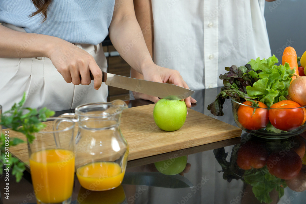 Closeup hand holding knife cutting green apple on a wooden chop board. Jar with mixed fruit juice and glass bowl with a variety of fruits and vegetables is placed on the kitchen counter. Closeup hand holding knife cutting green apple on a wooden chop board. Jar with mixed fruit juice and glass bowl with a variety of fruits and vegetables is placed on the kitchen counter.
