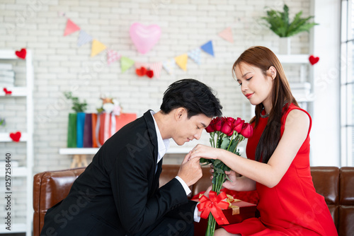 Asian man in a black suit kisses a woman hand while presenting her with a bouquet of red roses in a romantic scene setting for proposal of marriage