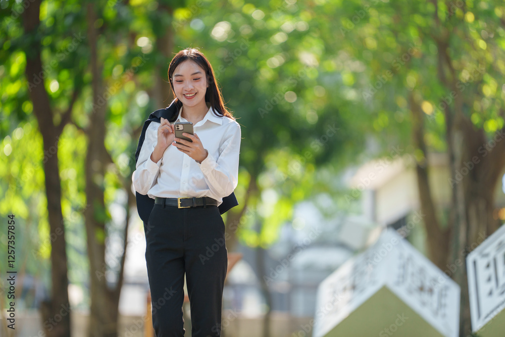Smiling young professional woman using a smartphone while walking in a sunny park, surrounded by lush greenery, conveying a sense of connectivity and modern lifestyle Smiling young professional woman using a smartphone while walking in a sunny park, surrounded by lush greenery, conveying a sense of connectivity and modern lifestyle