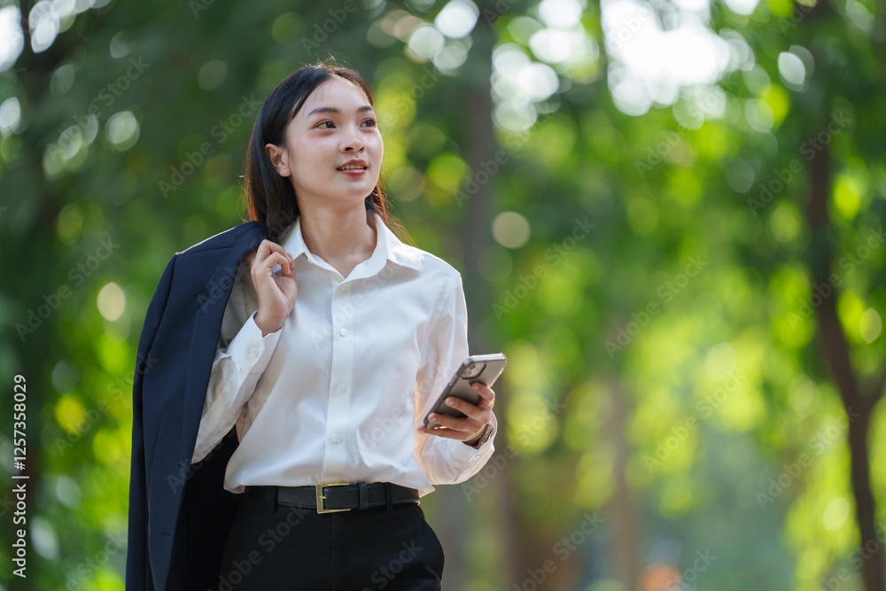 Businesswoman strolling through a lush park, holding a smartphone and a jacket. She is surrounded by greenery, exuding confidence and professionalism in a serene outdoor setting Businesswoman strolling through a lush park, holding a smartphone and a jacket. She is surrounded by greenery, exuding confidence and professionalism in a serene outdoor setting