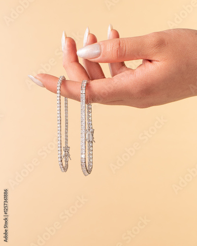 Elegant female hand with shimmering white manicured nails holding two sparkling diamond tennis bracelets against a soft peach background. Luxury jewelry concept.