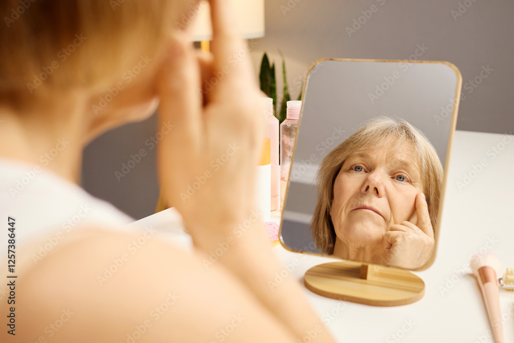 Naklejka premium A woman inspects her skin closely in a mirror while applying skincare products at home. The soft, natural light highlights her focused expression as she begins her morning routine