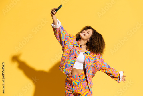 Happy young woman taking selfie through smart phone against yellow background