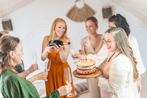 Birthday surprise party. Group of cheerful female friends celebrating personal success stories. Smiling girls toasting with glasses of champagne one holding a birthday cake with candles to blow. photo