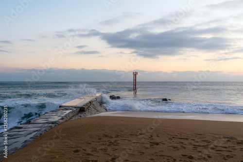 Les vagues déferlent sur la jetée de la Barre d'Étel, projetant des embruns sur les roches sombres. Sous un ciel nuageux, l’océan impose sa force dans ce paysage sauvage et contrasté.