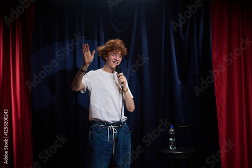 Performer delivering stand-up comedy on stage with microphone in hand, engaging audience with humor and gestures, red curtains framing stage photo