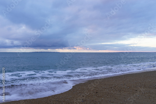 Coucher de soleil sur la Barre d’Étel sous un ciel nuageux teinté de rose. La mer calme lèche le sable doré, laissant une fine écume éphémère sur le rivage. Une atmosphère douce et apaisante.