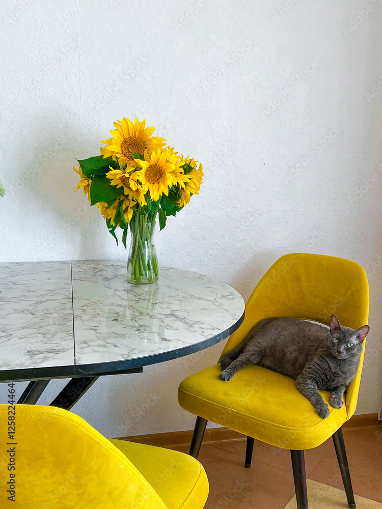 A grey cat is lying on a yellow armchair near a table with a bouquet of sunflowers