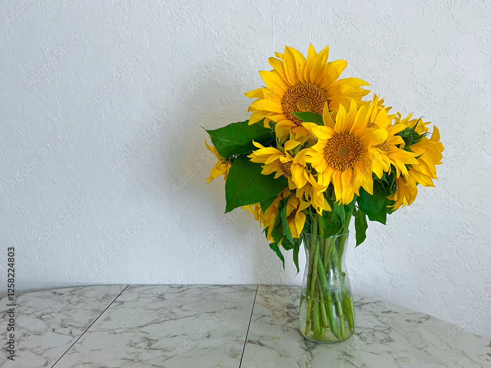 A bouquet of sunflowers stands in a glass vase on a marble table in a white kitchen