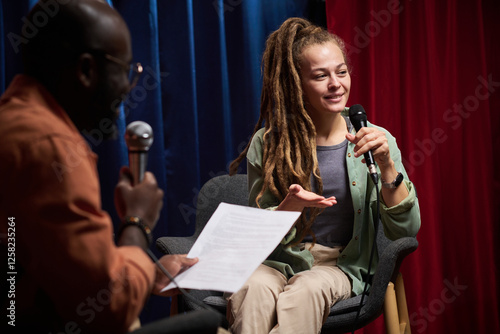 Canvas Print Smiling woman engaged in conversation while sitting in chair holding microphone, with interviewer nearby holding notes