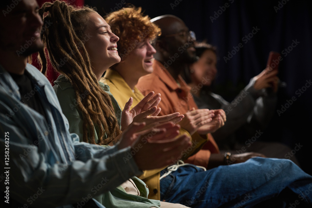 Obraz premium Group of diverse individuals of various ethnicities enjoying and applauding a performance, seated closely together in a darkened room sharing collective experience and connection