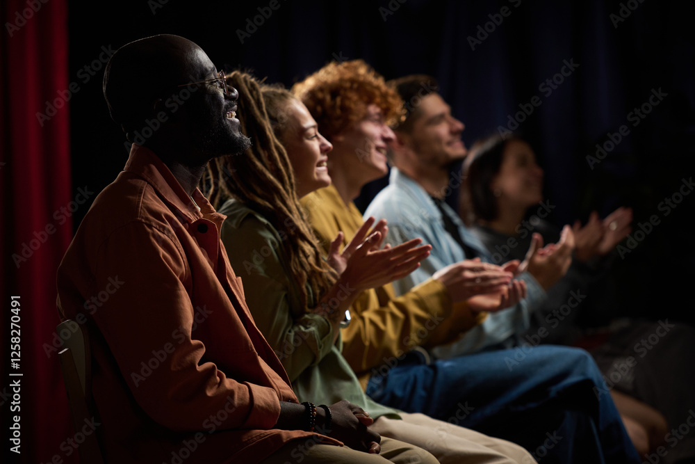 Obraz premium Group of diverse individuals applauding a performance with joyful expressions while seated in a dimly lit environment, highlighting togetherness and enjoyment