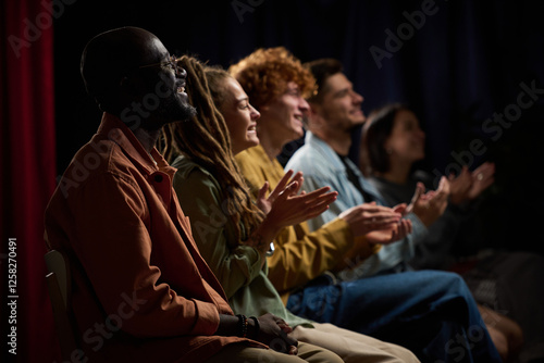 Group of diverse individuals applauding a performance with joyful expressions while seated in a dimly lit environment, highlighting togetherness and enjoyment